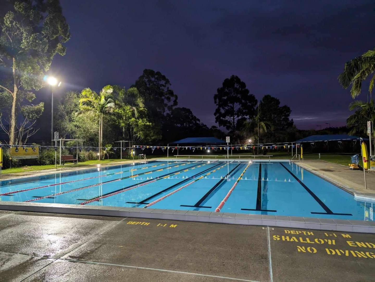 The view of the 25m outdoor heated pool at Tilligerry Aquatic Centre at night. No one is in the pool and the water is calm. A light is shining above the calm pool.