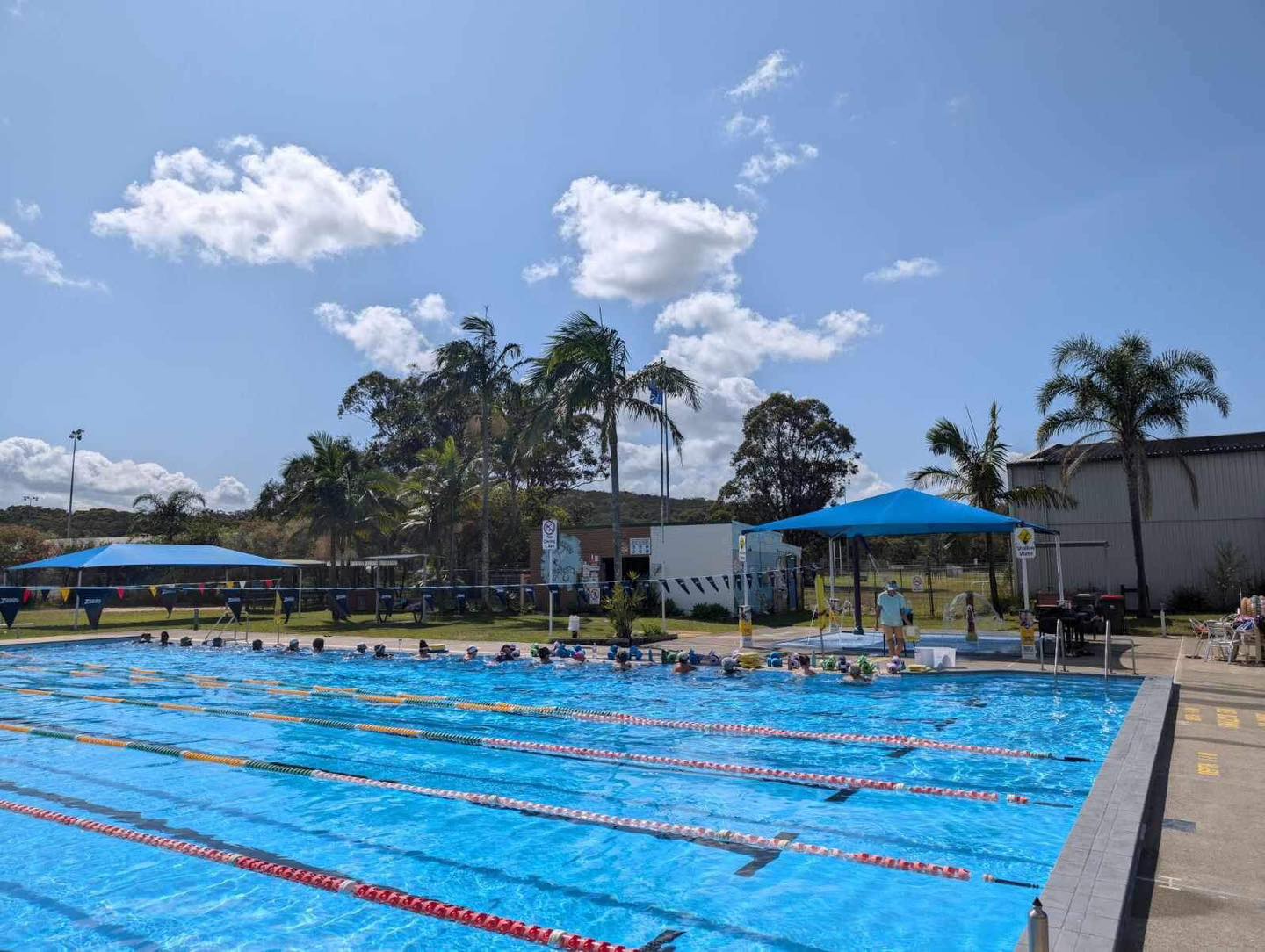 Featured is the Tilligerry Aquatic Centre 25m outdoor pool on a sunny day with blue skies and a few white clouds. The water appears still. Some people are participating in a class and resting in the water on the edge of the pool.