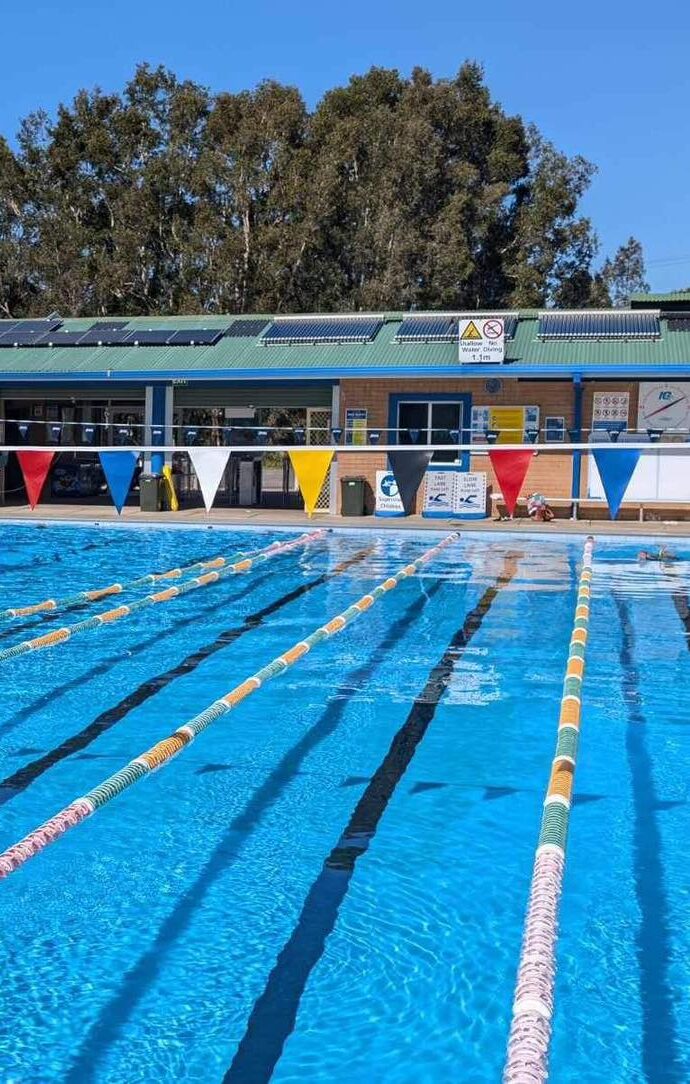 Featured is the Tilligerry Aquatic Centre's 25m outdoor heated pool. There are yellow, blue, white and red triangle bunting hanging above the pool on a blue sky day.